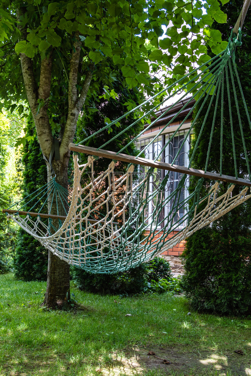 柳条吊床，固定在树干上，在常绿植物和乡村别墅的模糊背景下。特写镜头。庆祝的气氛。阳光明媚的一天。宁静的休息和放松。照片摄影图片