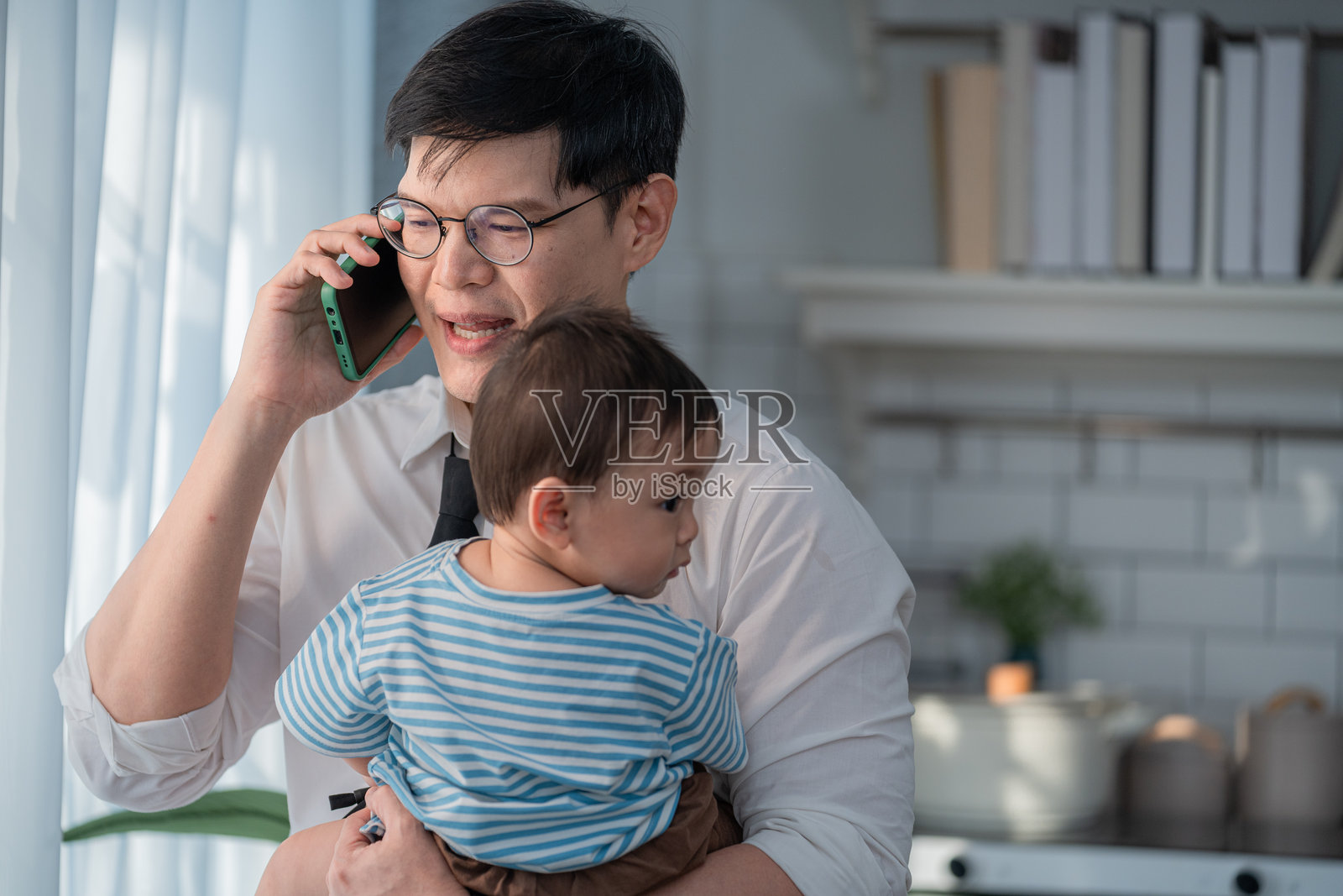 父亲在明亮的家居环境中一边打电话一边抱着他的幼儿照片摄影图片