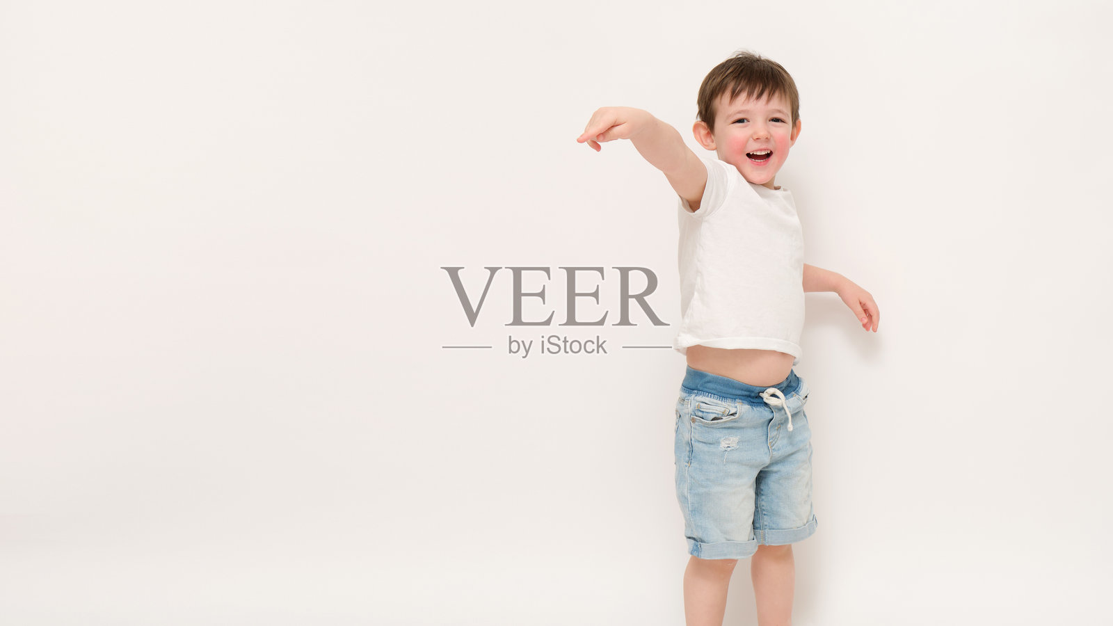 微笑的男孩穿着休闲夏季服装，指向一侧。白色背景下的工作室肖像。儿童生活方式和顽皮童年概念。三岁的小孩。照片摄影图片