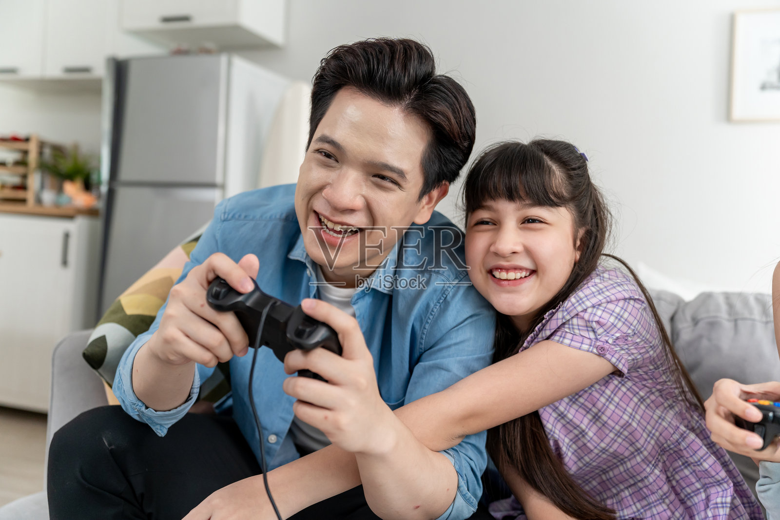 亚洲父母与年轻女儿在家中玩游戏机。迷人的父亲和小女孩坐在沙发上，感到兴奋，享受在家客厅里一起玩游戏的时光。照片摄影图片