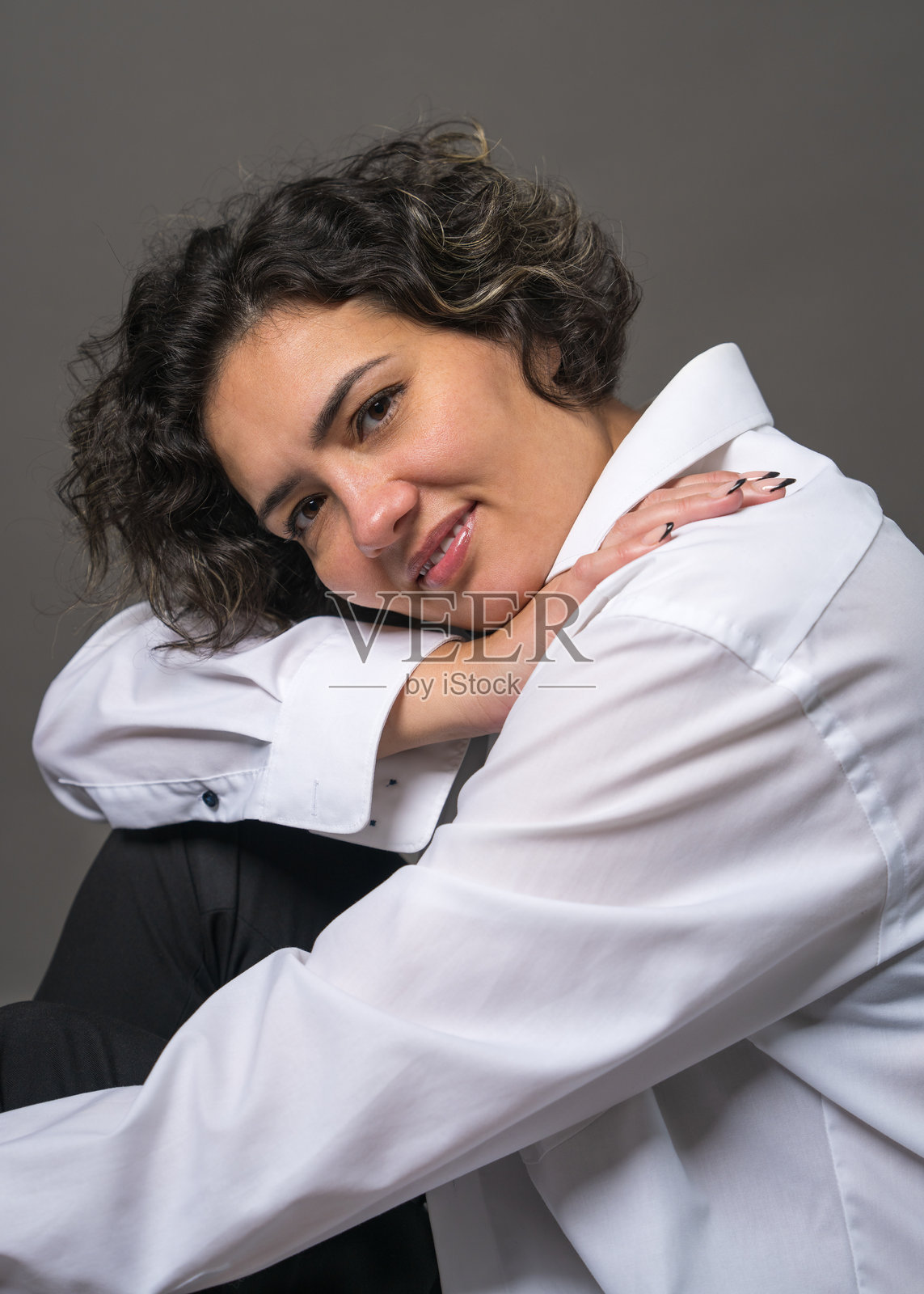 优雅迷人的年轻深发女性，穿着白衬衫，坐在地板上，头低垂在双手上。背景为灰色，女性模特在摄影棚中摆姿势。照片摄影图片