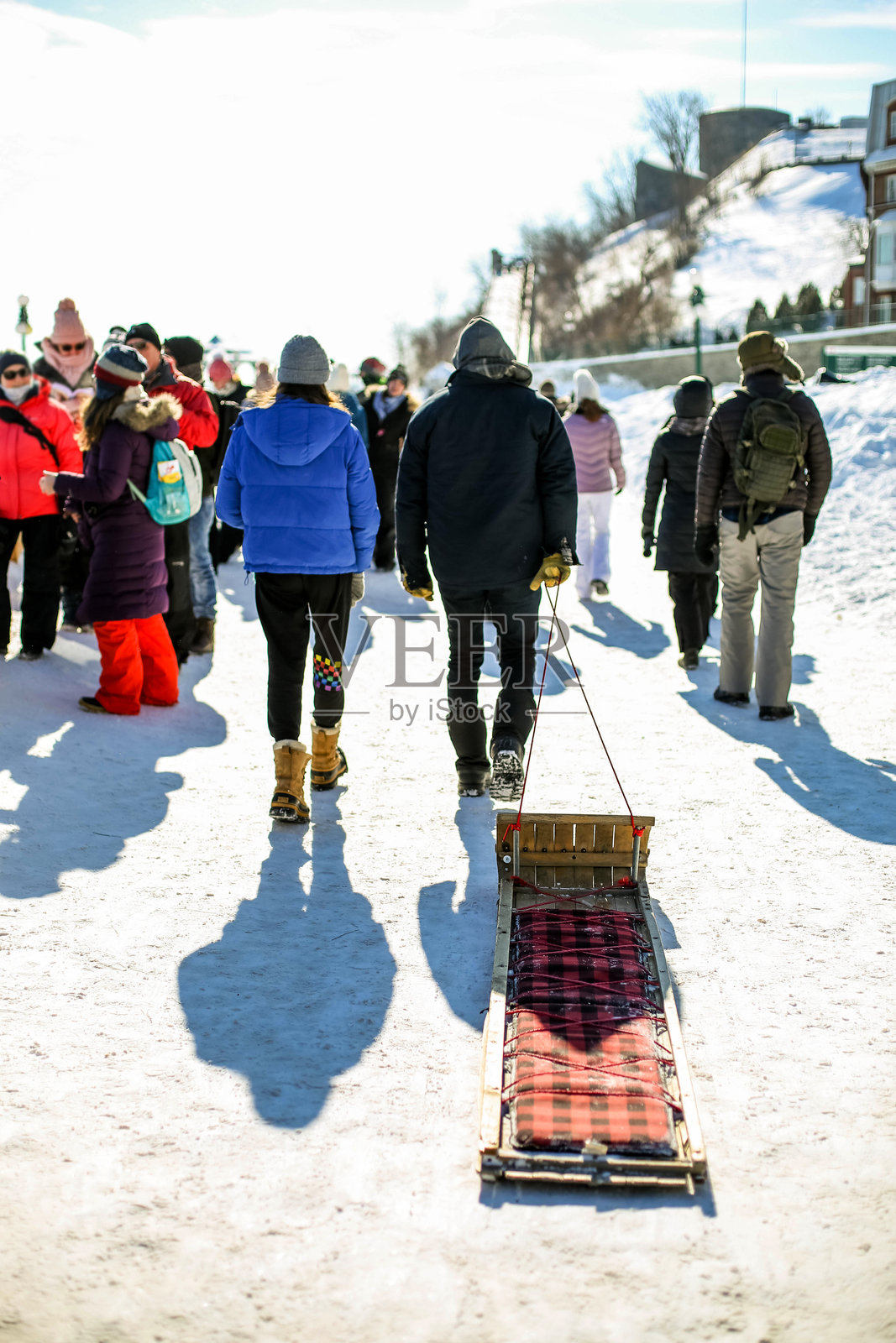 男子在冬季魁北克嘉年华的拥挤街道上拉着雪橇朝冰滑梯走去照片摄影图片