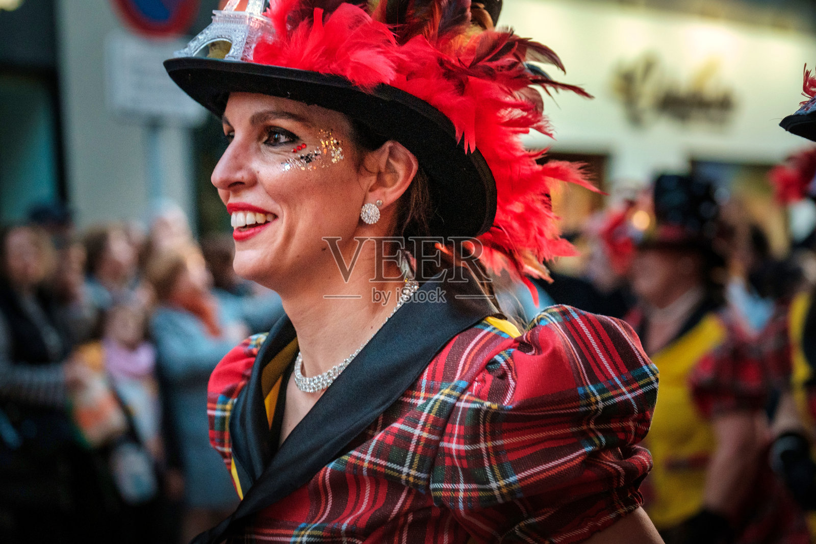 穿着节日服装和配饰的女性,包括一件格子衣物和一顶装饰有红色羽毛的帽子,脸部化妆,眼周有装饰性点缀,可能是作为服装的一部分或为了特别场合。照片摄影图片