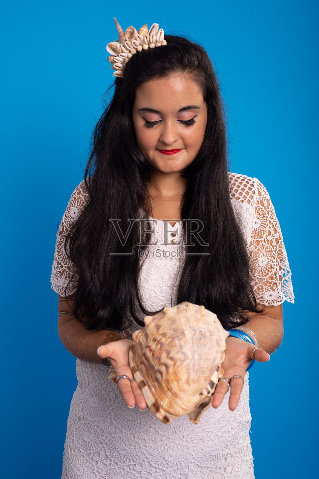 美丽的年轻女子穿着白色连衣裙，头上戴着贝壳头饰，手里拿着贝壳，看着贝壳。照片摄影图片