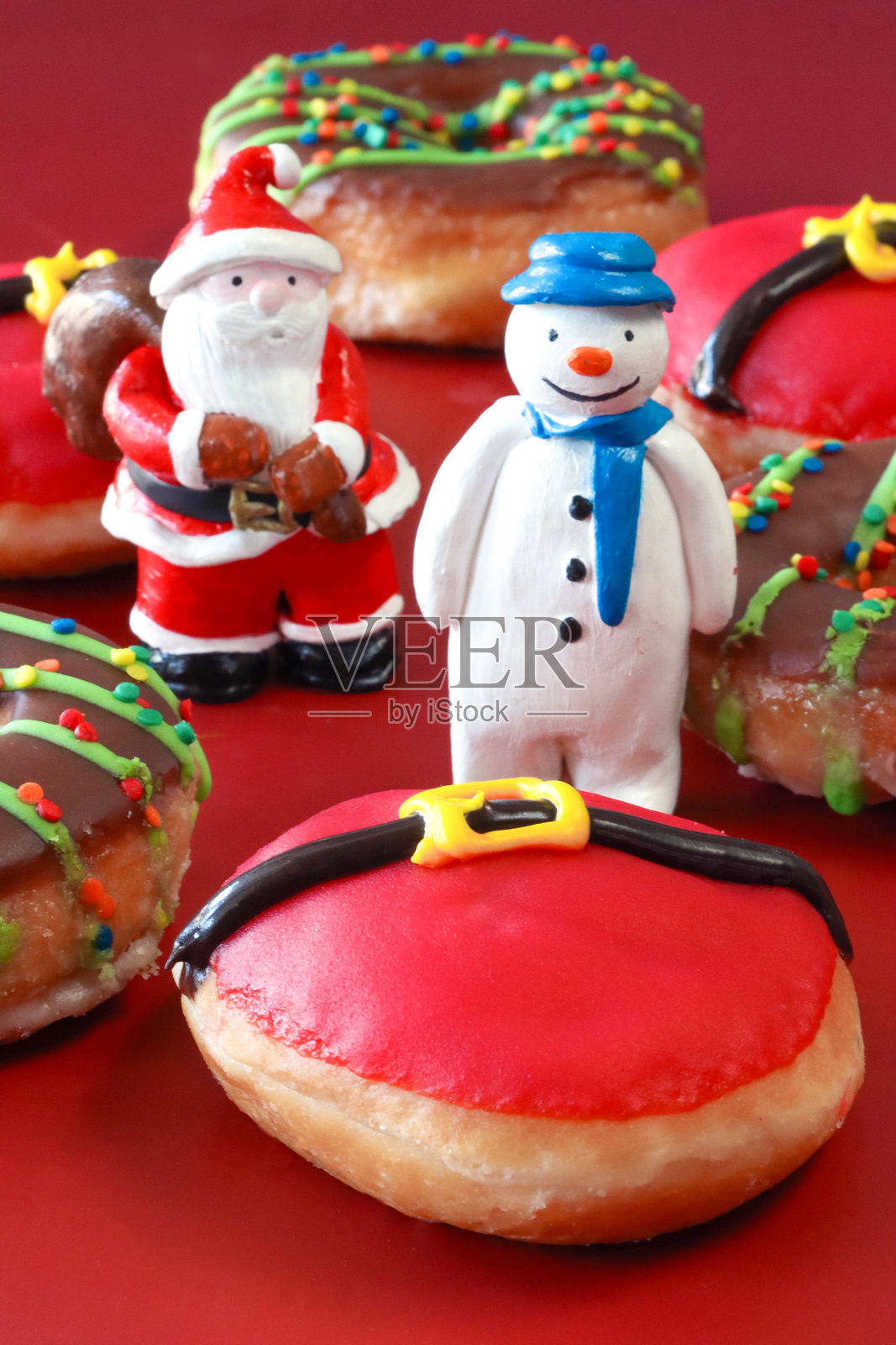 圣诞老人和雪人小雕像的特写图像与一批圣诞老人肚皮糖冰甜甜圈和巧克力釉面环甜甜圈，油炸面团甜点装饰红色糖霜和扣带细节，红色背景，重点在前景照片摄影图片