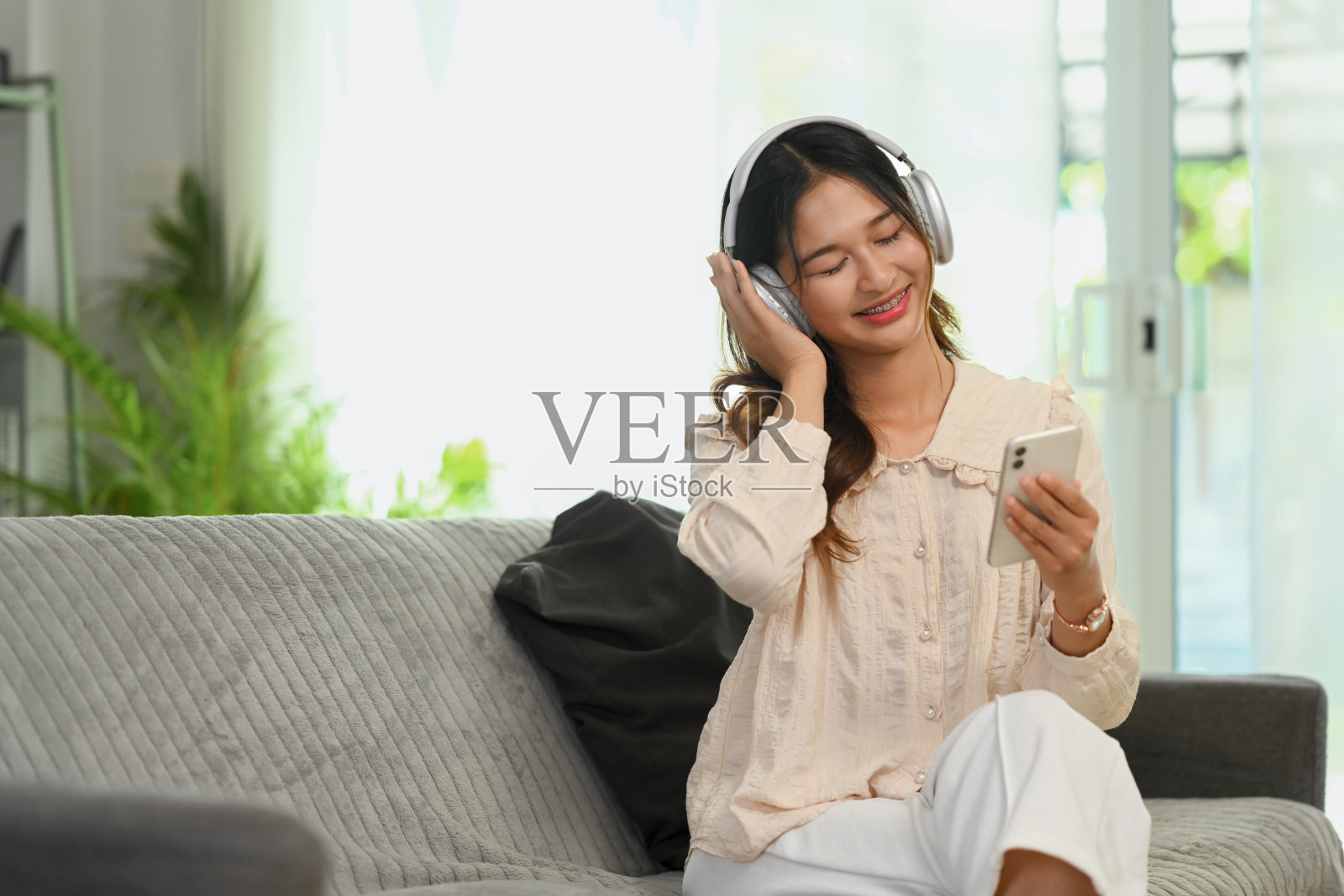 开朗的年轻女子戴着耳机听音乐，在沙发上使用智能手机。照片摄影图片