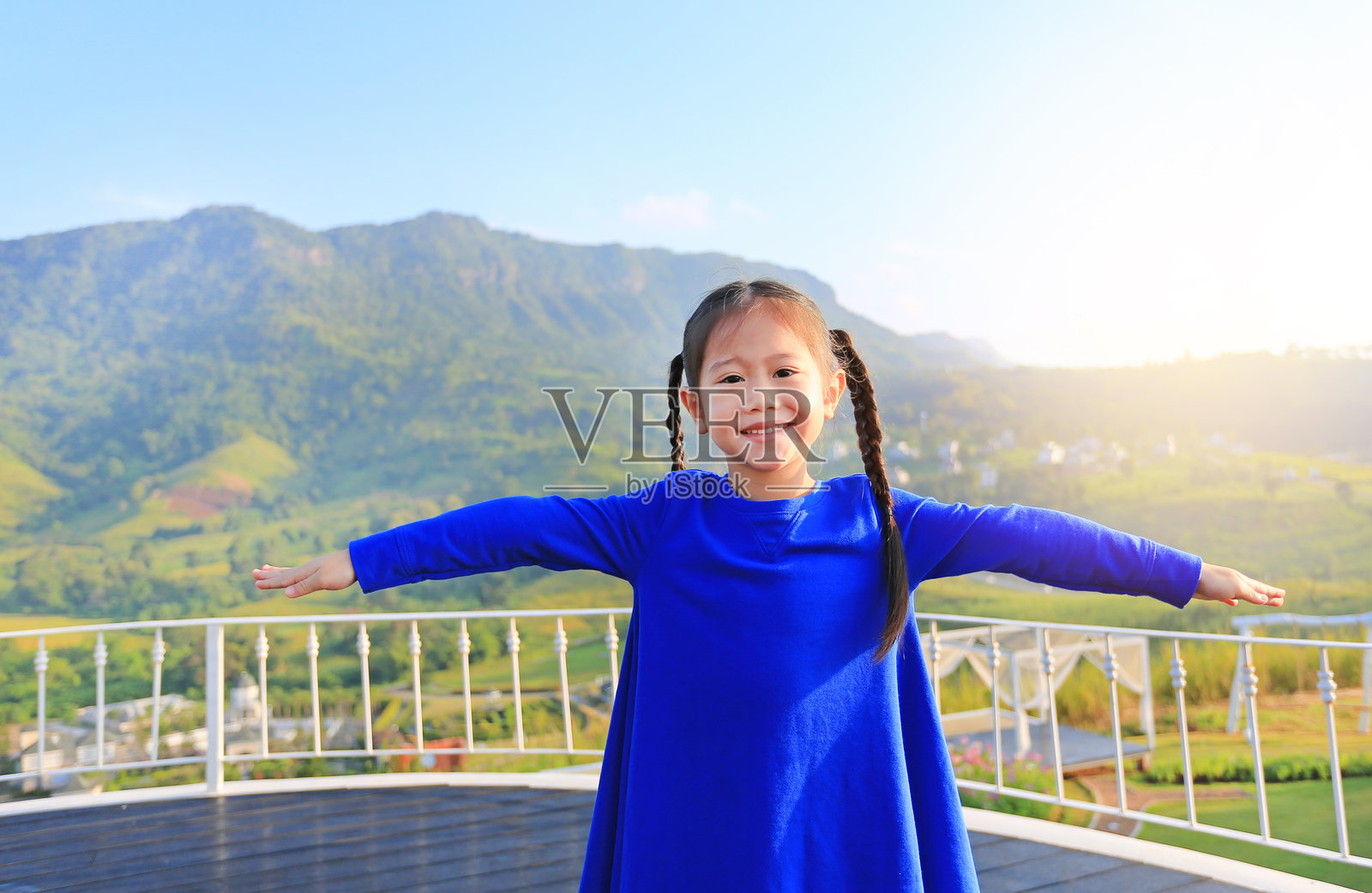 可爱的亚洲小女孩在蓝天上张开双臂，看着美丽的树木和山脉，感觉很自由。照片摄影图片
