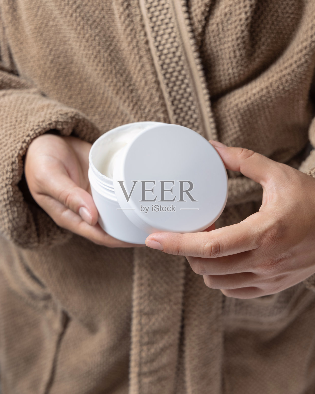 穿着棕色浴袍的女性特写，正在用手打开白色护肤霜罐，化妆品样品展示照片摄影图片