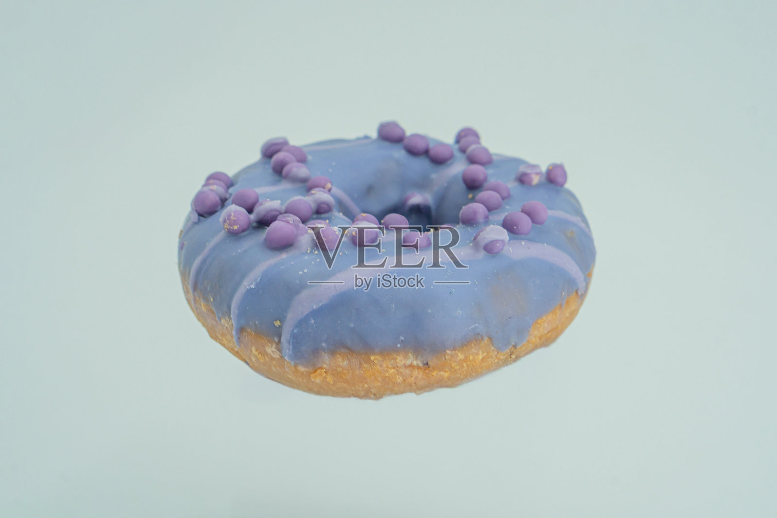 一个色彩鲜艳的单个甜甜圈，底部呈金棕色，上面涂有薰衣草色的糖霜，并点缀有紫色、蓝色和白色的装饰，放置在浅色背景上。图像展示了糕点的形状和质感。照片摄影图片