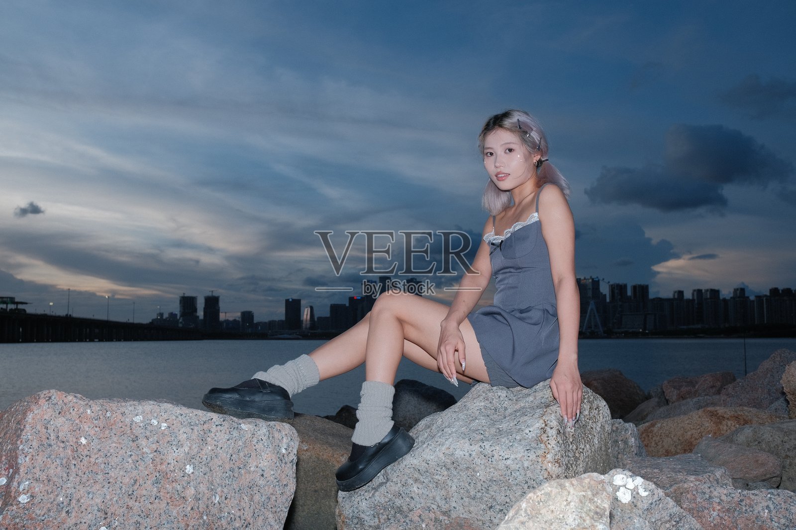 年轻女性坐在海边的岩石上，背对着夕阳的天空。照片摄影图片