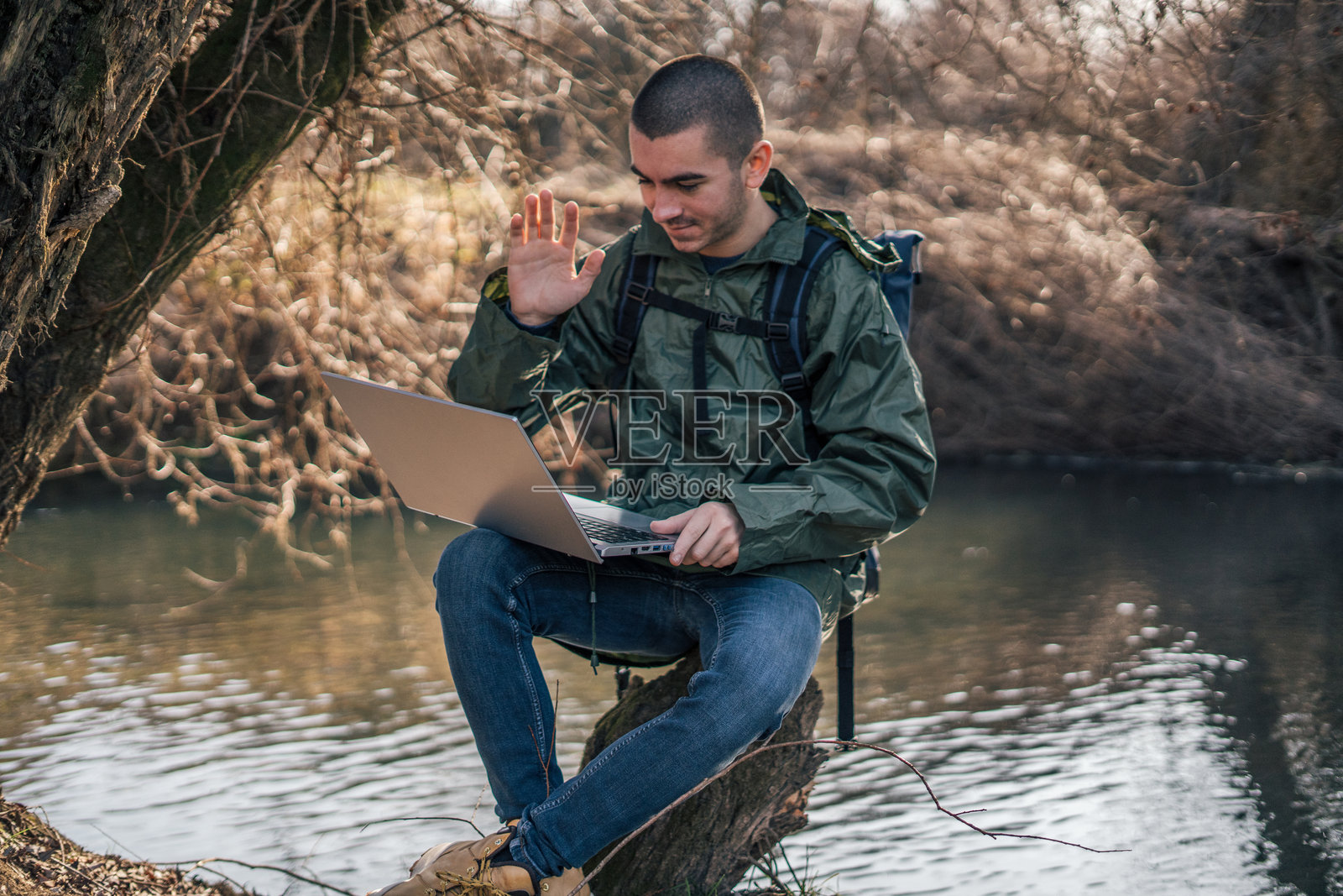 一位年轻帅气的徒步旅行者在大自然中放松，背着一个大背包，使用笔记本电脑，愉快地向视频通话中的朋友挥手。照片摄影图片