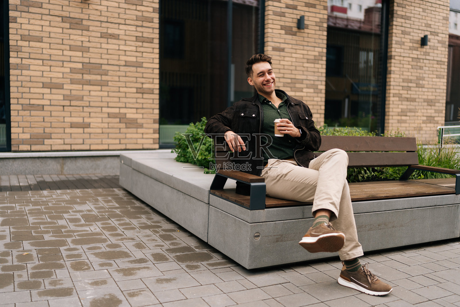 阳光明媚的秋日，一位快乐的年轻人坐在城市长椅上，靠在砖砌建筑旁，品味着咖啡，享受着城市环境的愉悦氛围。照片摄影图片