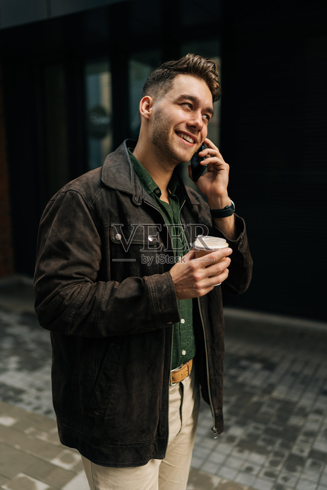 穿着休闲服的快乐男子的竖版肖像，手持外卖咖啡杯，在现代建筑外的城市环境中进行电话交谈。照片摄影图片