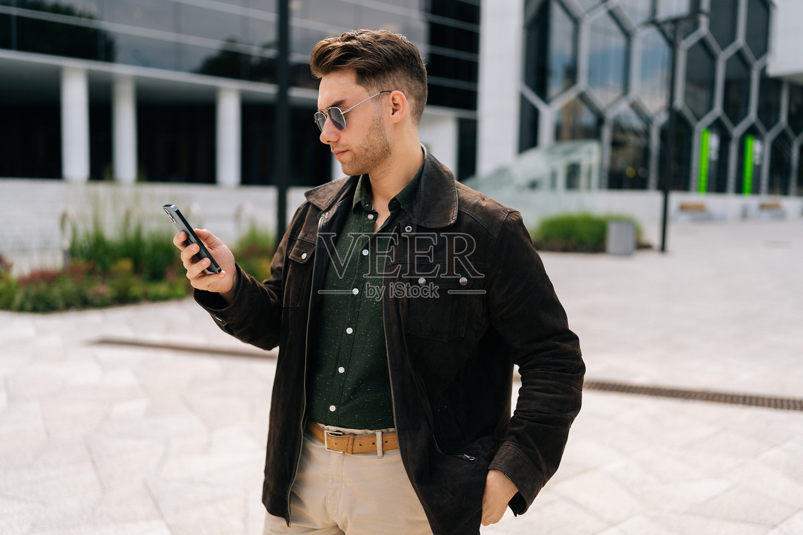 时尚的商人戴着太阳镜，手持手机，在城市街道上走动时浏览互联网，阅读信息，靠近现代办公大楼。照片摄影图片