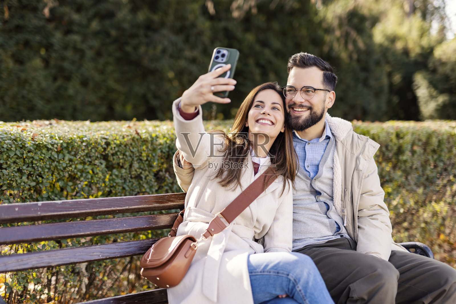 年轻情侣坐在城市公园的长椅上，拥抱并拍自拍。照片摄影图片