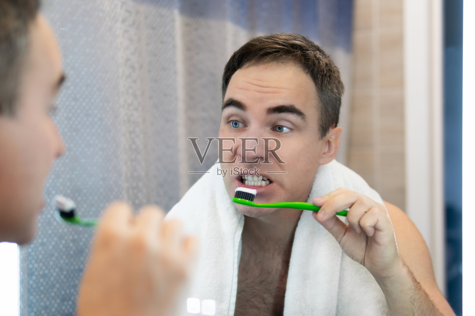 年轻男子在家浴室的镜子前刷牙，镜中的他正在刷白牙。照片摄影图片