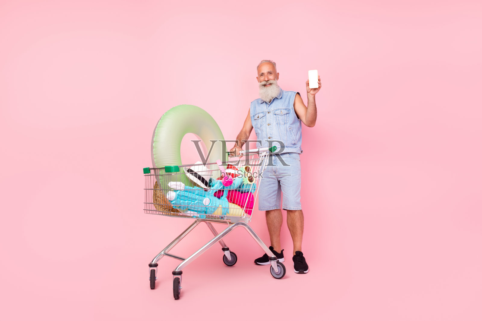 穿着牛仔裤和马甲的酷退休男士全身照，走在购买夏季装备的空旷空间，背景为粉色。照片摄影图片