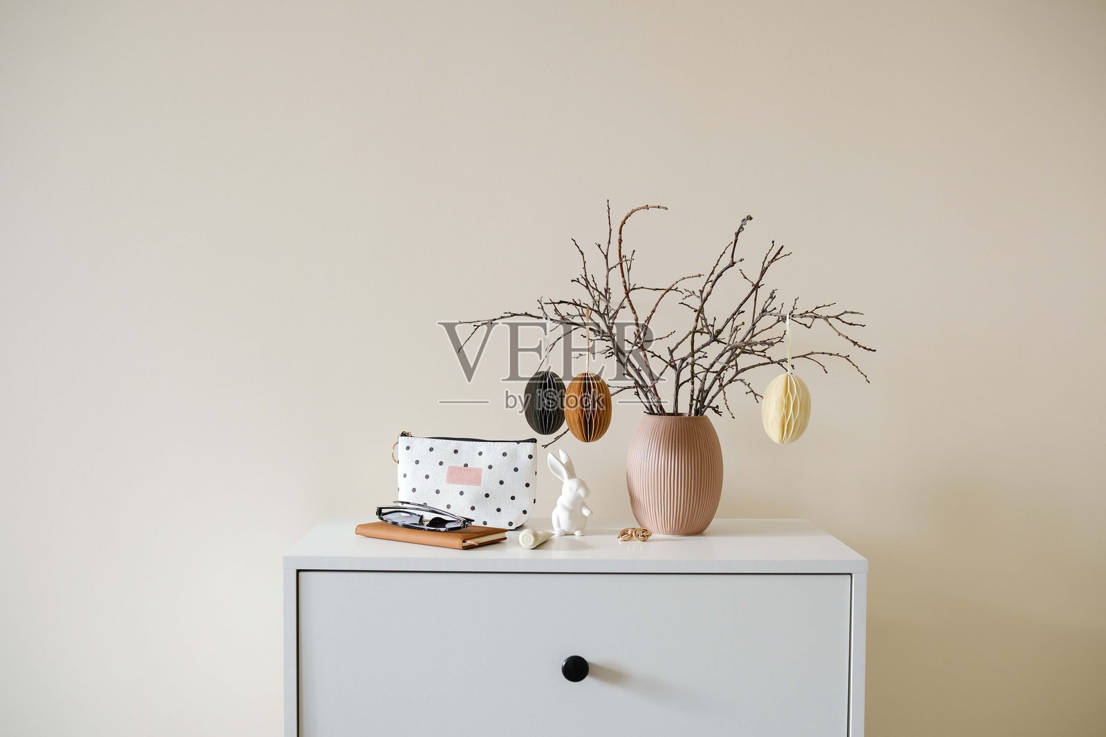 时尚简约的复活节装饰，干枝上优雅地排列着手工彩蛋，放置在现代梳妆台上的简单花瓶中，梳妆台上还有女性配饰 - 笔记本和化妆包。照片摄影图片