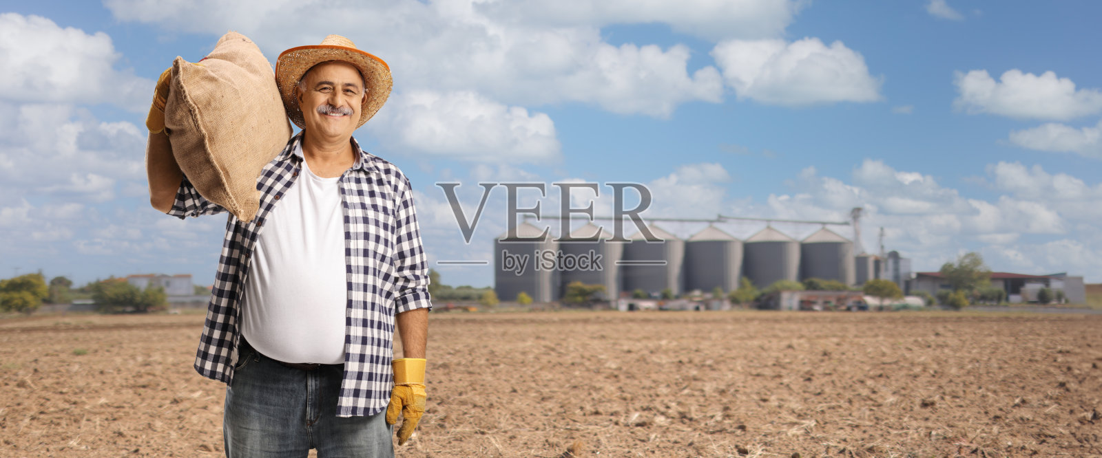 成熟的农民肩上扛着麻布袋，站在有塔式粮仓的田野上照片摄影图片