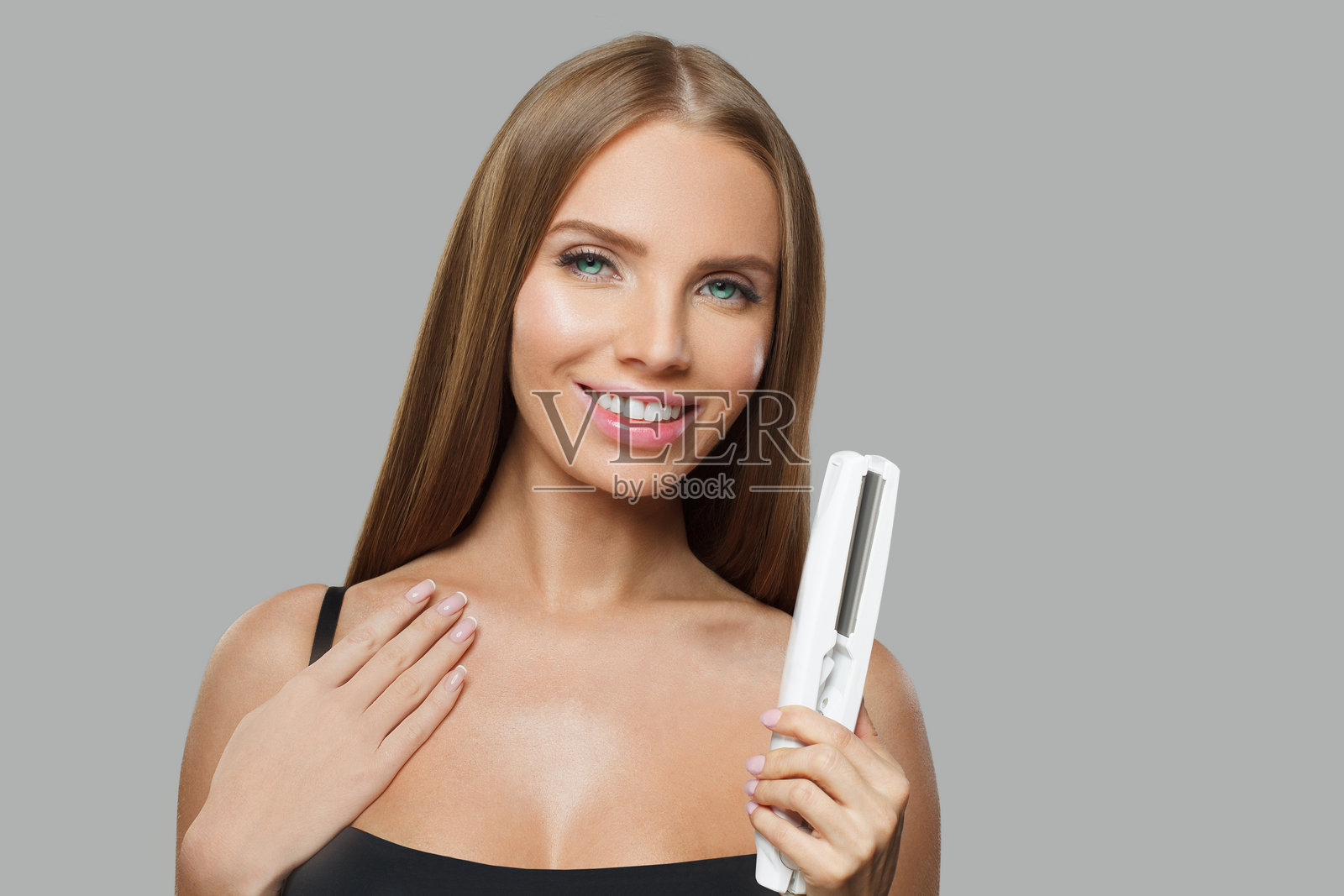 快乐的时尚模特，棕色光滑的直发肖像。可爱的女性手持直发器，背景为灰色摄影棚。照片摄影图片