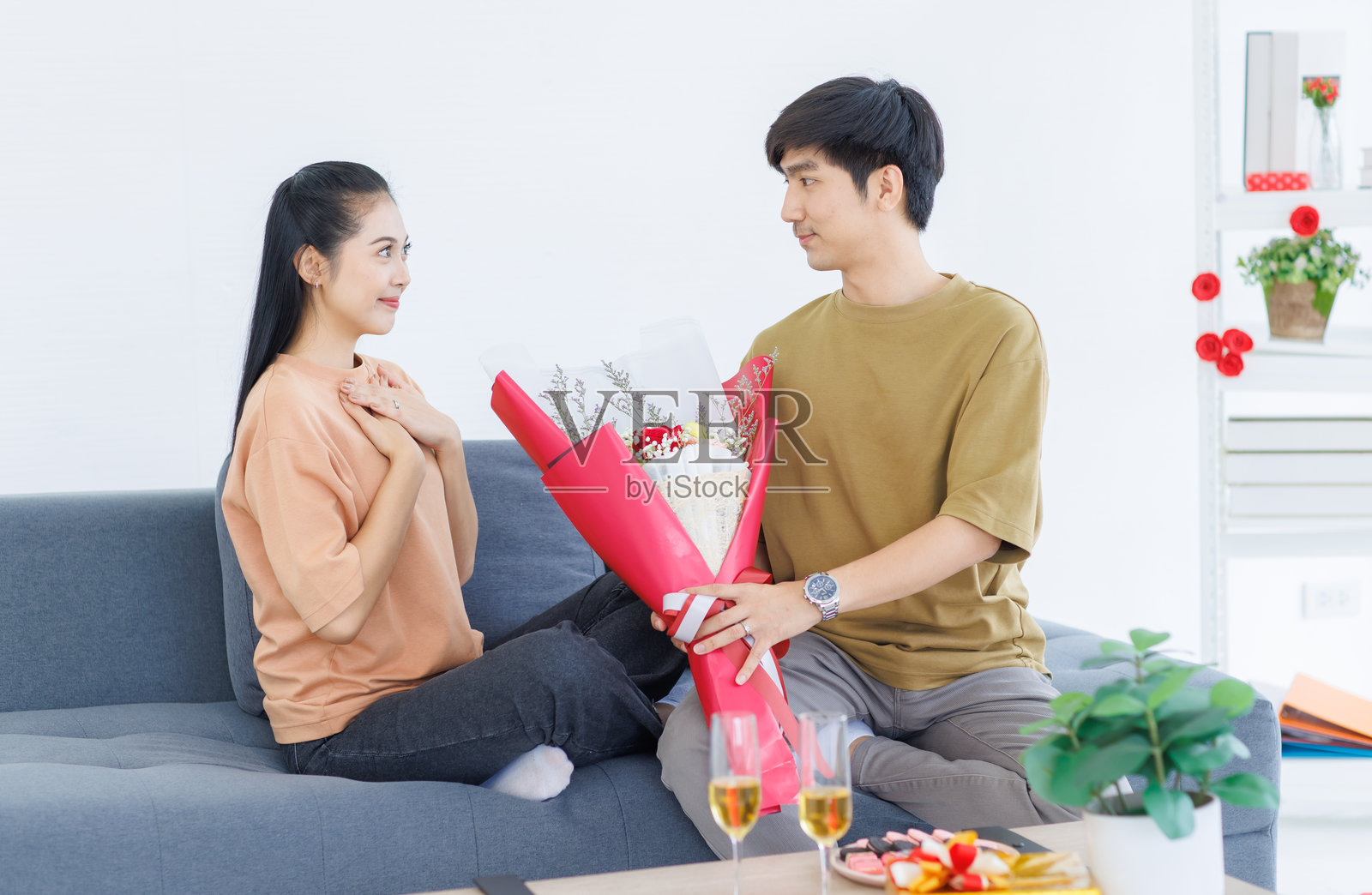 亚洲年轻快乐迷人的情侣，甜蜜的微笑，给对方惊喜的红玫瑰花束，坐在家中舒适的沙发上，享受情人节的浪漫时光。照片摄影图片