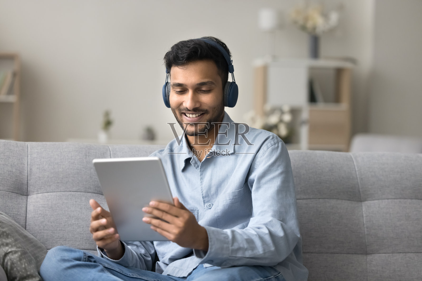 印度男子戴着耳机坐在沙发上，手里拿着平板电脑照片摄影图片