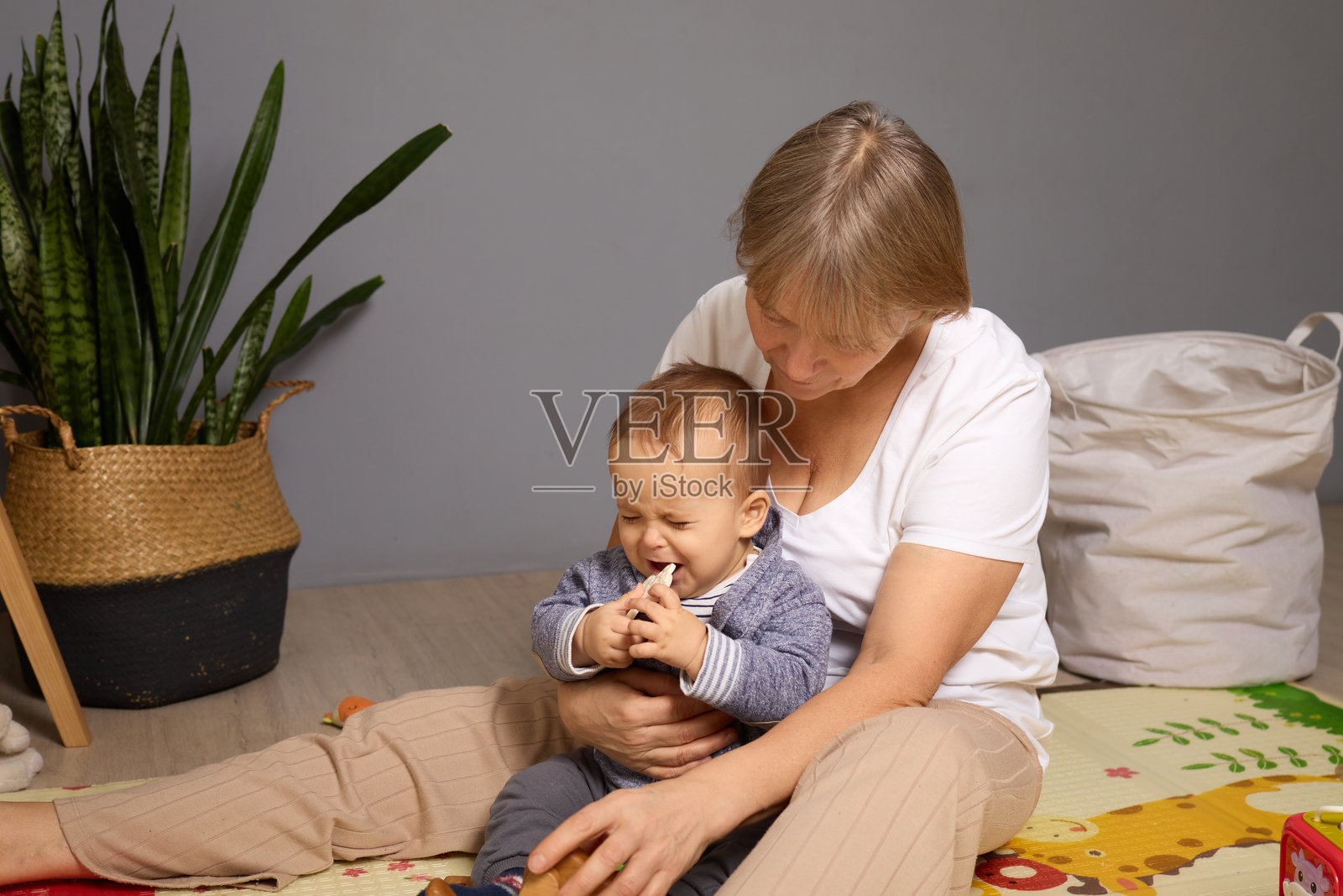 一位女性坐在地板上，和一个男婴一起微笑，享受着玩耍的时光。房间里有植物和活泼的装饰，营造出温馨的氛围。照片摄影图片