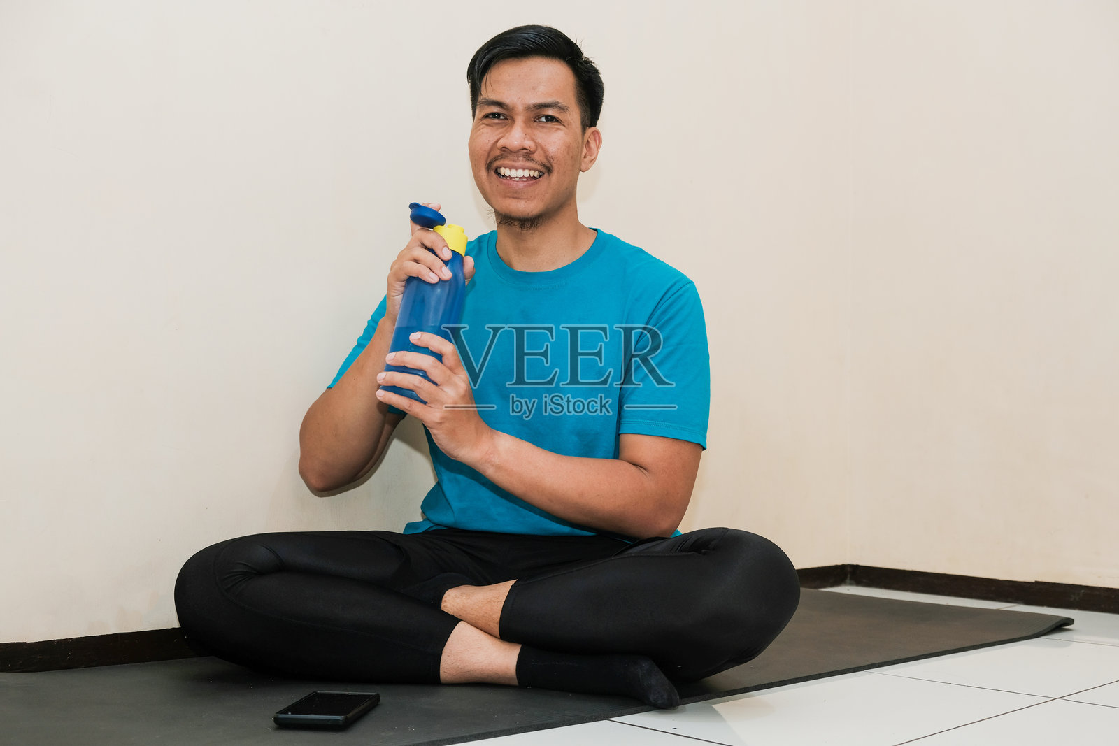 微笑的东南亚男子快乐地坐在健身垫上，手里拿着蓝色水瓶，背景为纯色奶油色。照片摄影图片