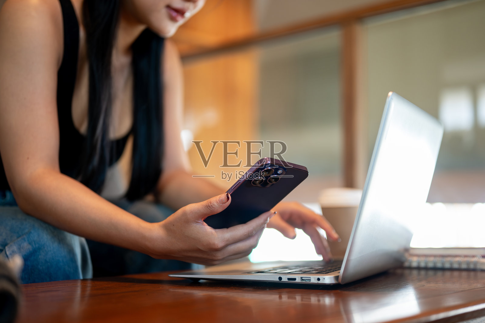一位亚洲女性在咖啡店里使用手机，同时远程工作于她的笔记本电脑。照片摄影图片