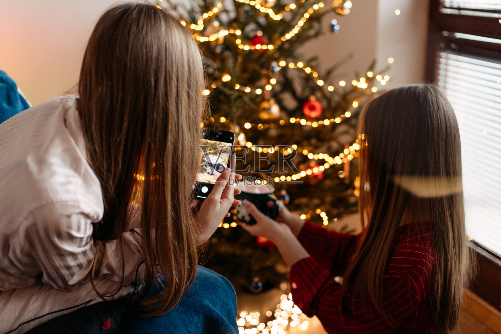两个青少年女孩姐妹在家中圣诞树旁，温馨的氛围中用杯子装着棉花糖的可可互相拍照。照片摄影图片