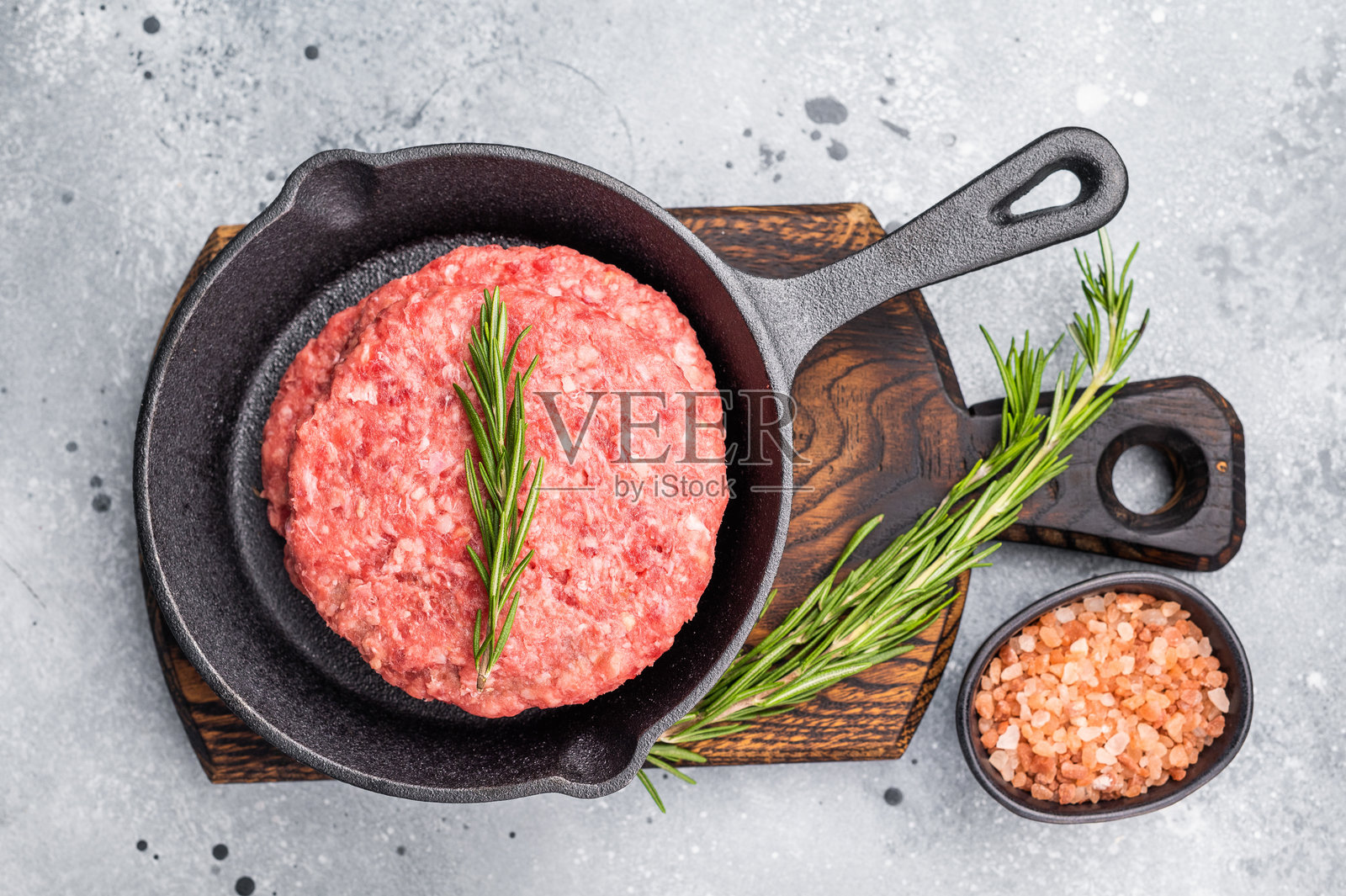 准备好烹饪的生牛肉饼，带香草的炸肉饼在平底锅中。灰色背景。俯视图。照片摄影图片