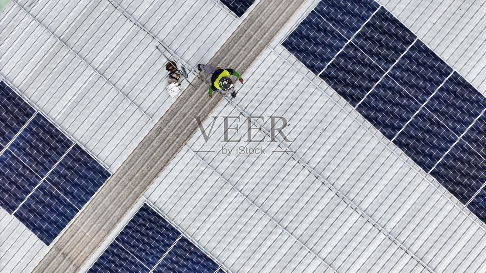 一名穿着安全装备的技术人员在金属屋顶上检查太阳能电池板，手中拿着工具。场景强调可再生能源、太阳能技术和可持续基础设施的维护。照片摄影图片