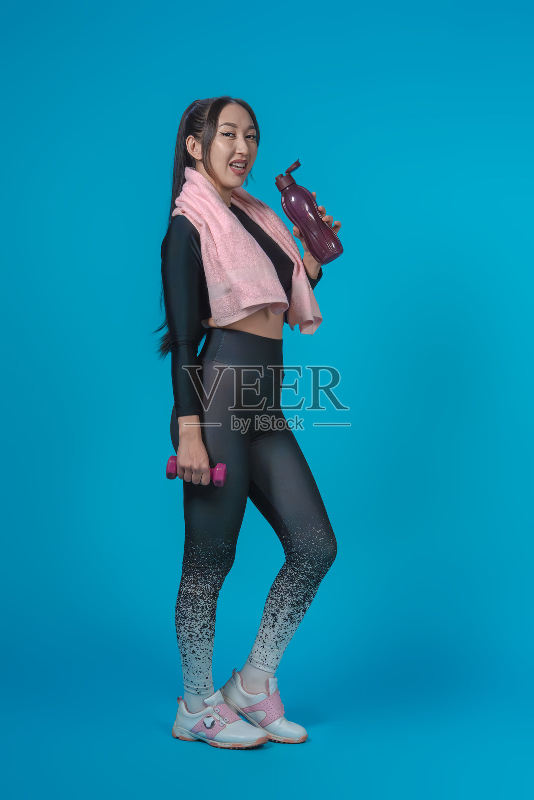 穿着运动服的瘦小亚洲女孩，脖子上挂着毛巾，手里拿着水瓶，刚刚锻炼完。背景为蓝色，健康生活方式的概念。照片摄影图片