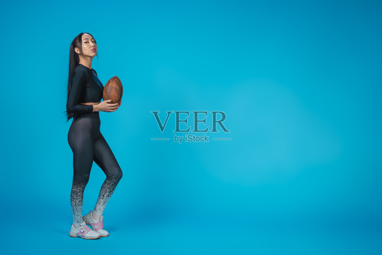 身材苗条的亚洲女孩穿着运动服，手持橄榄球，背景为纯蓝色。留白空间。照片摄影图片
