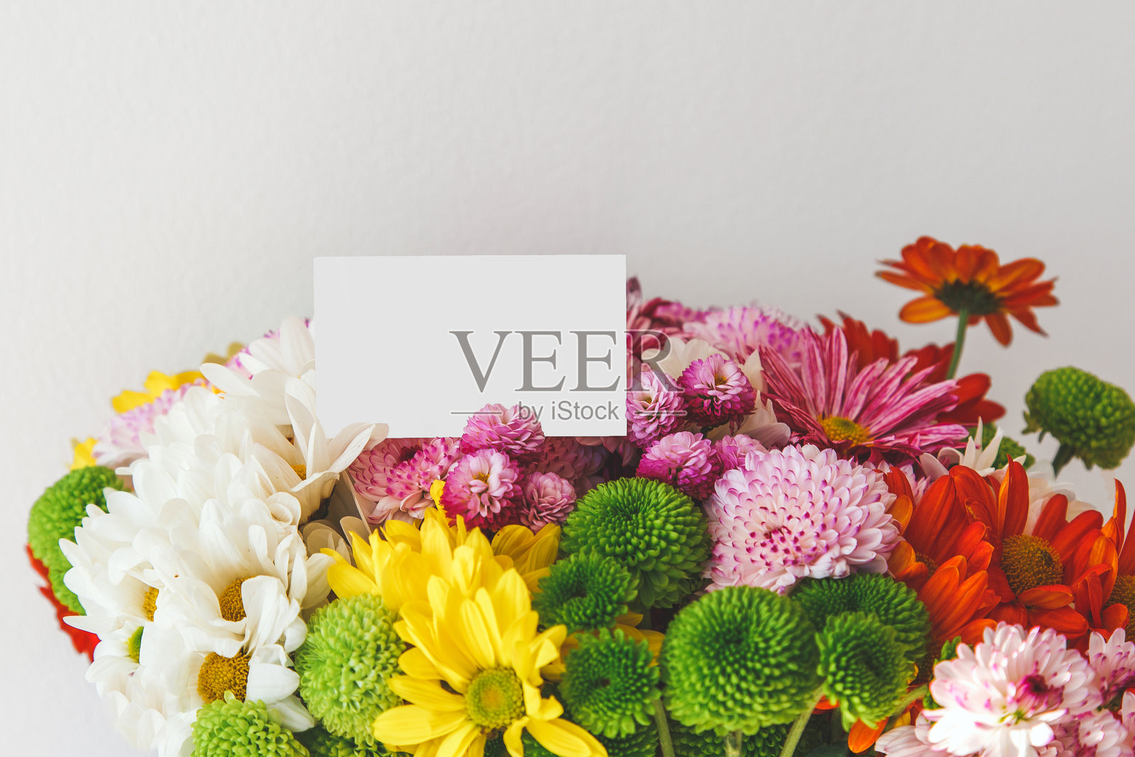 色彩缤纷的新鲜花束，搭配一张空白贺卡，包含白色、黄色、粉色、绿色和橙色的雏菊和菊花，背景为中性色，留有空白空间，样机。照片摄影图片