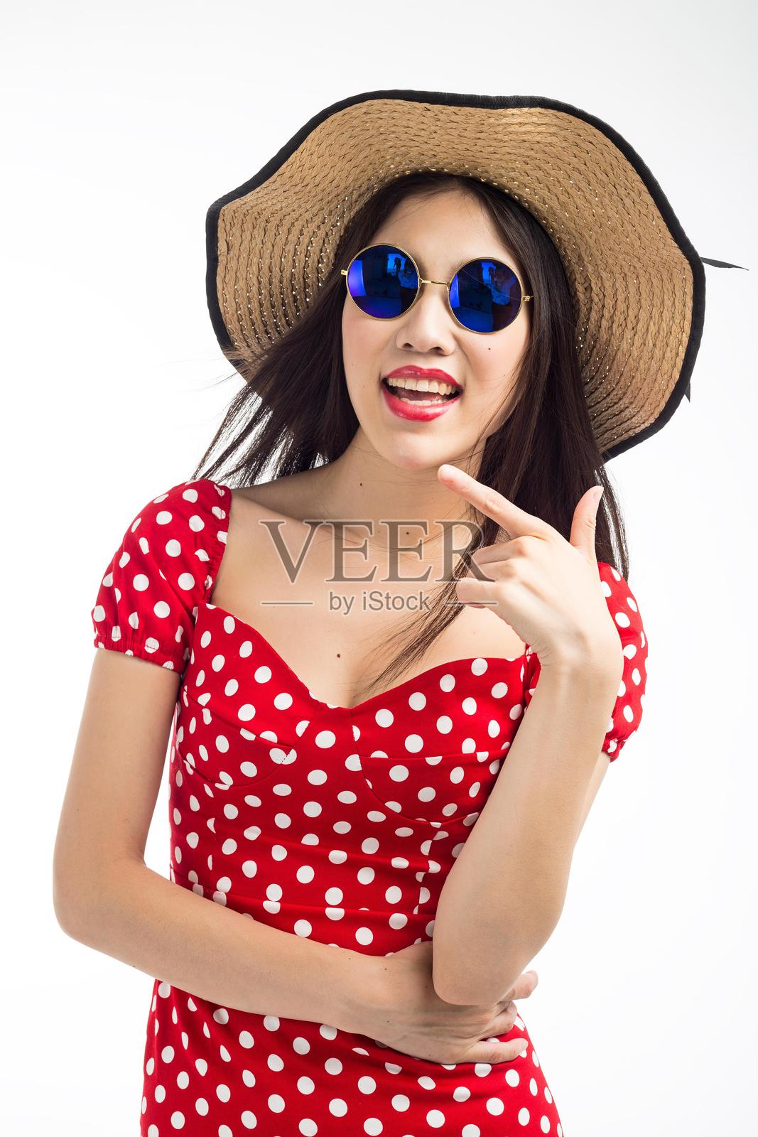 年轻的亚洲女性穿着红色波点裙，戴着太阳镜和草帽，伸出手指指向前方。照片摄影图片