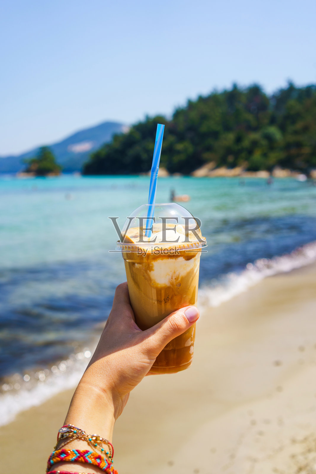 女人手持并展示一杯冰奶昔，面朝大海，阳光明媚的日子，视角特写照片摄影图片
