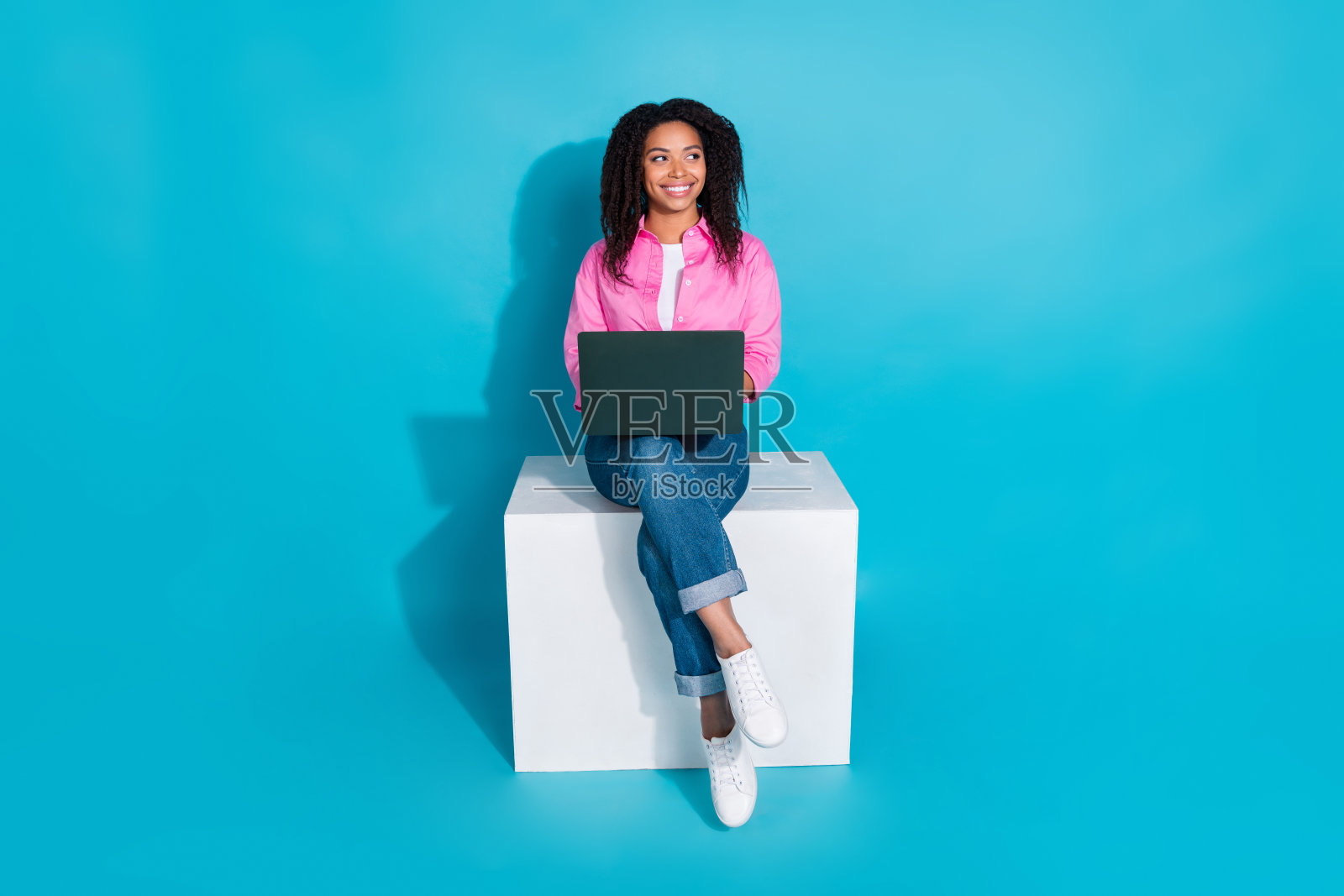 穿着粉色衬衫的年轻女性坐在立方体上，手持笔记本电脑，背景为蓝色，展现现代时尚风格。照片摄影图片