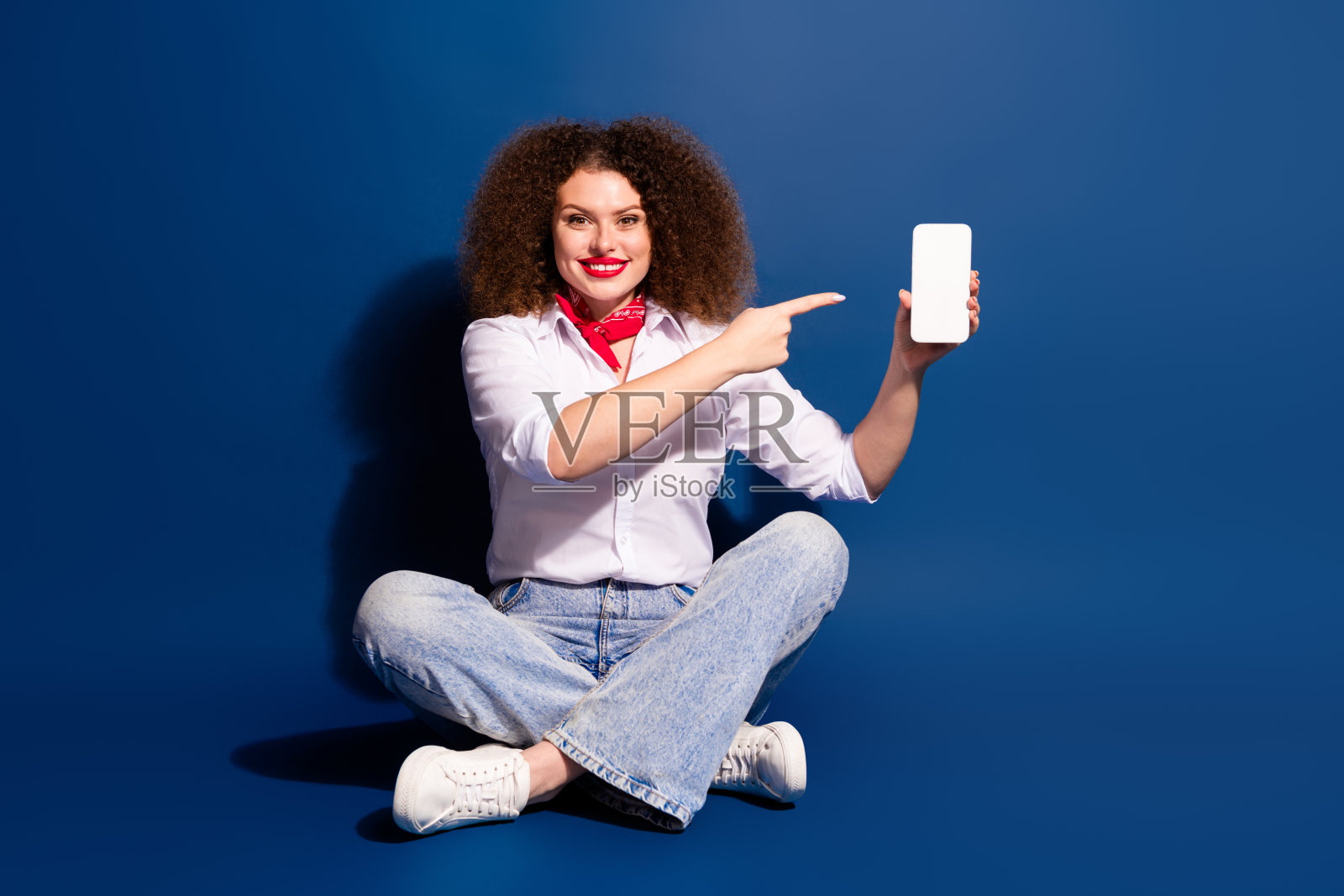 全身照，光鲜迷人的女士穿着红色手帕，指着苹果iPhone和三星现代电子产品，背景为空白的蓝色。照片摄影图片