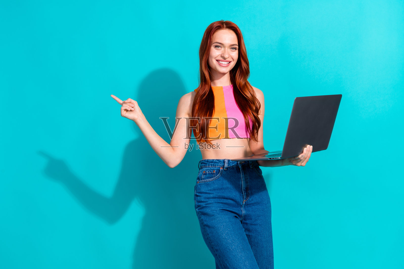 微笑的年轻红发女性穿着休闲服装，指着青绿色背景上的笔记本电脑照片摄影图片