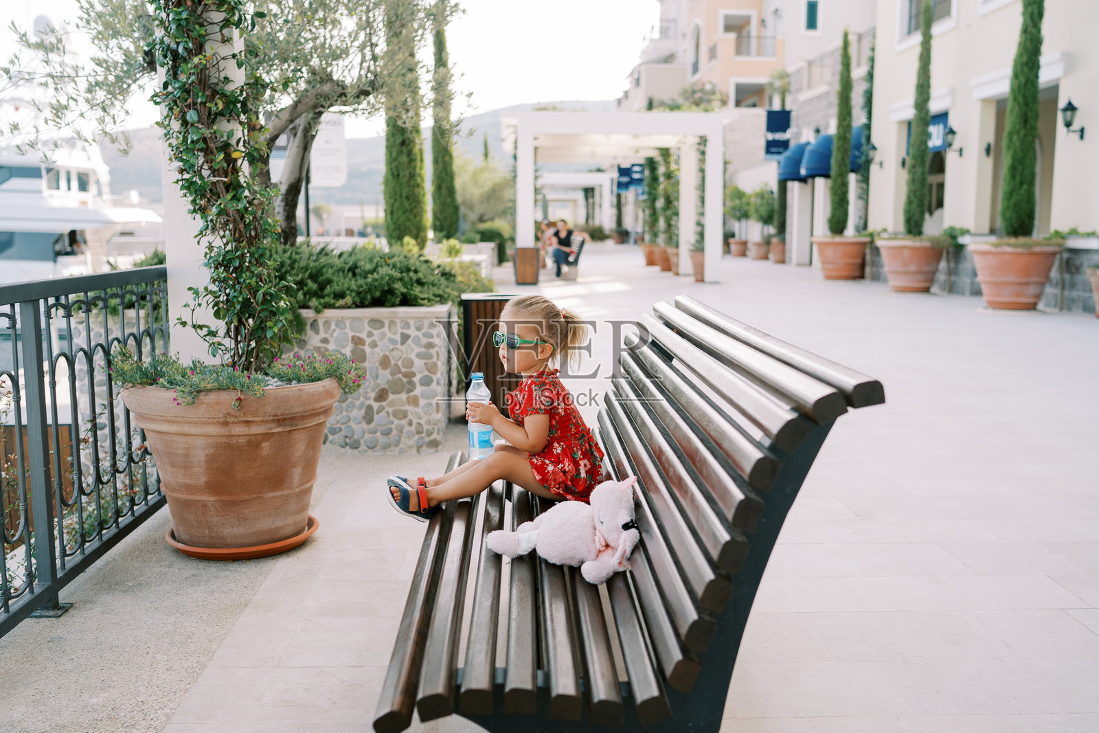 小女孩手里拿着一瓶水，坐在海滨长廊的长椅上，侧身望着大海。照片摄影图片