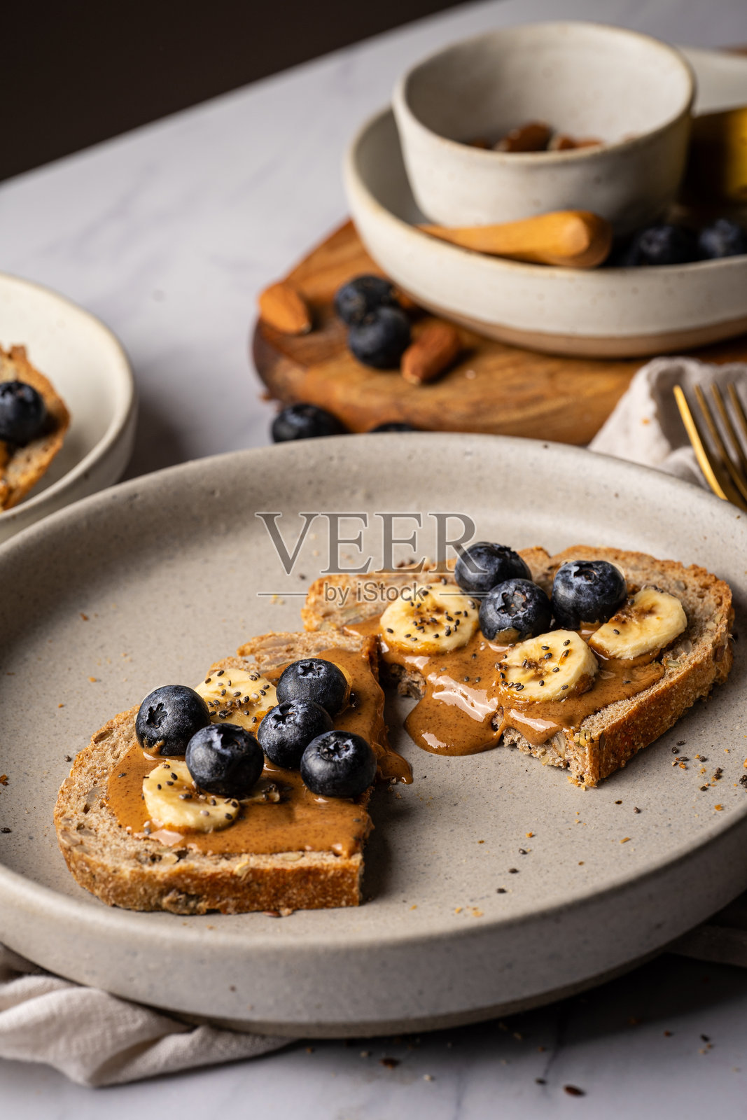 酸面包吐司，上面涂有杏仁酱，撒上蓝莓、香蕉和奇亚籽，摆放在桌子上。杏仁酱正在滴落。健康的甜美早餐。照片摄影图片