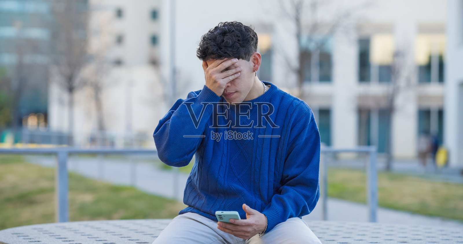 年轻的拉丁美洲男子坐着，手里拿着智能手机，面露失望，手遮住脸。照片摄影图片