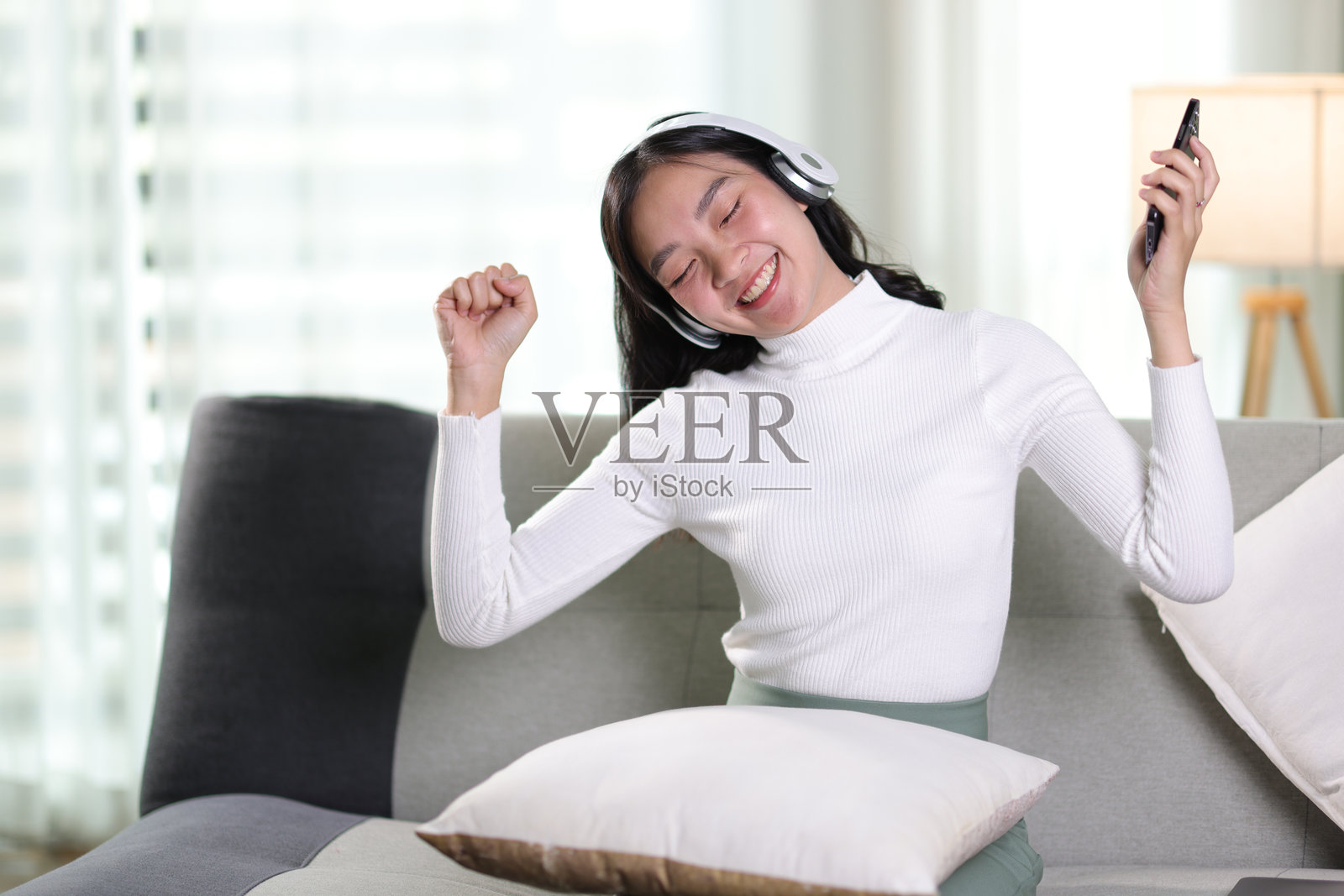 年轻的亚洲女性戴着耳机坐在家里的沙发上使用智能手机。快乐的女孩在客厅放松舒适，度过慵懒的周末，享受听音乐，看电影。照片摄影图片