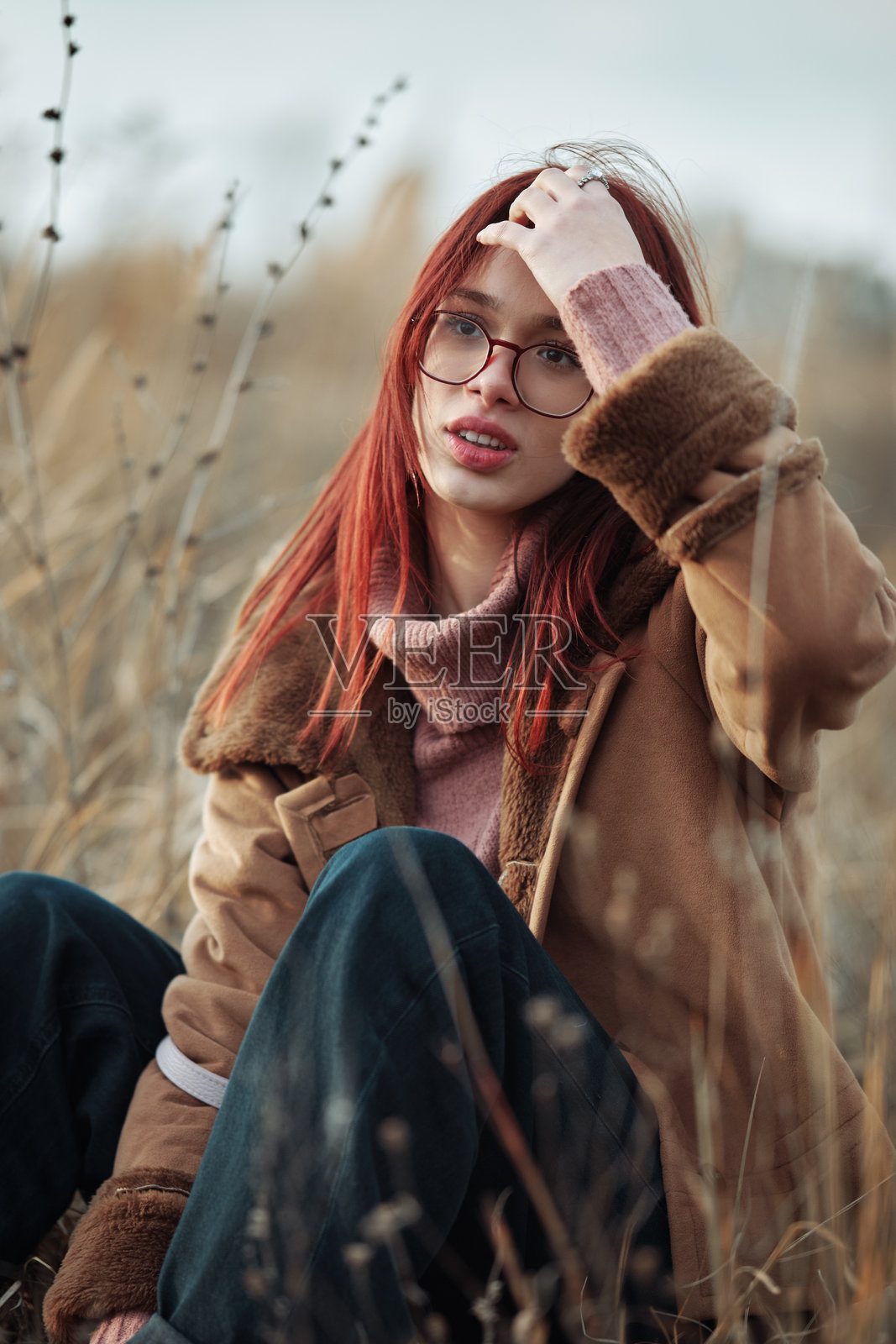 红发女孩戴着眼镜，坐在高高的草丛中，手指轻触头发，神情若有所思，穿着温暖，迎着秋风。照片摄影图片