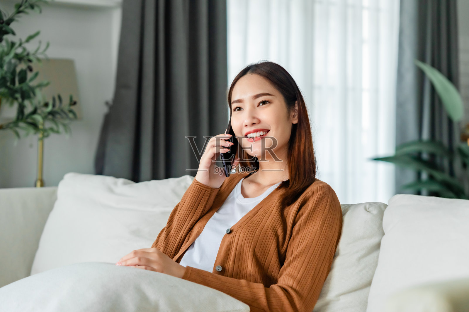 快乐的年轻亚洲女性在家中舒适的沙发上放松，微笑着用智能手机发短信聊天。照片摄影图片