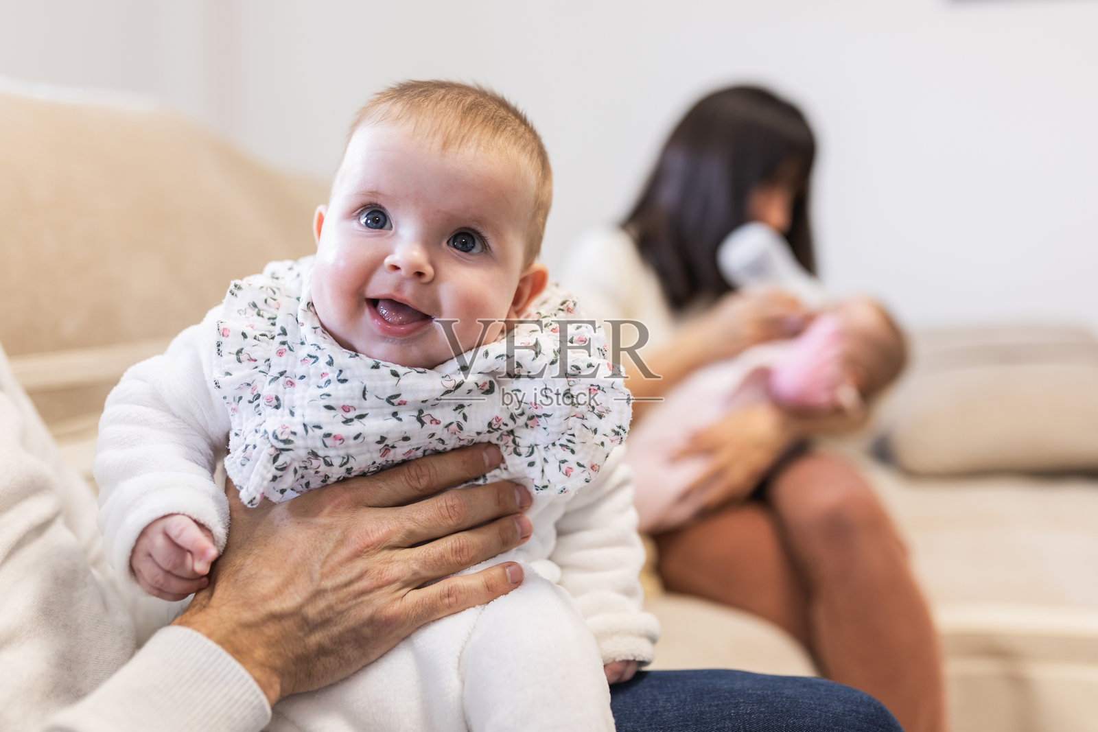 母亲坐在沙发上，抱着一个婴儿双胞胎，正在用奶瓶喂养他们，父亲坐在旁边，陪伴着另一个双胞胎。照片摄影图片