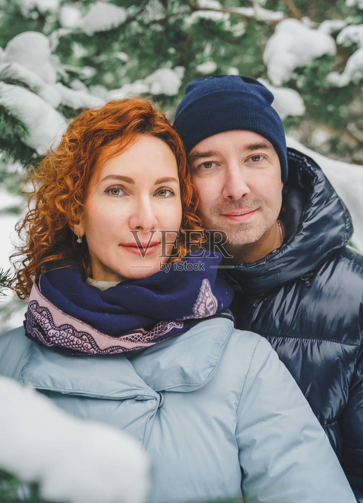浪漫爱情故事。快乐的中年千禧夫妇，冬季雪地公园里一位红发女子。浪漫约会，亲吻，拥抱。散步，享受乐趣。时尚的蓝色衣服，夹克，帽子。照片摄影图片
