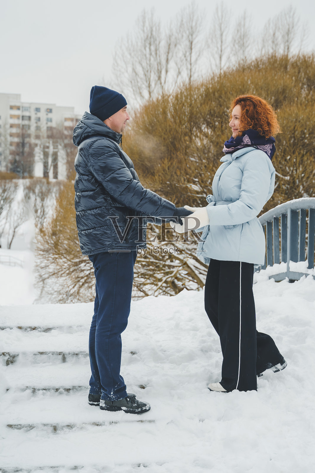 浪漫爱情故事。快乐的中年千禧夫妇，冬季雪地公园里一位红发女子。浪漫约会，亲吻，拥抱。散步，享受乐趣。时尚的蓝色衣服，夹克，帽子。照片摄影图片