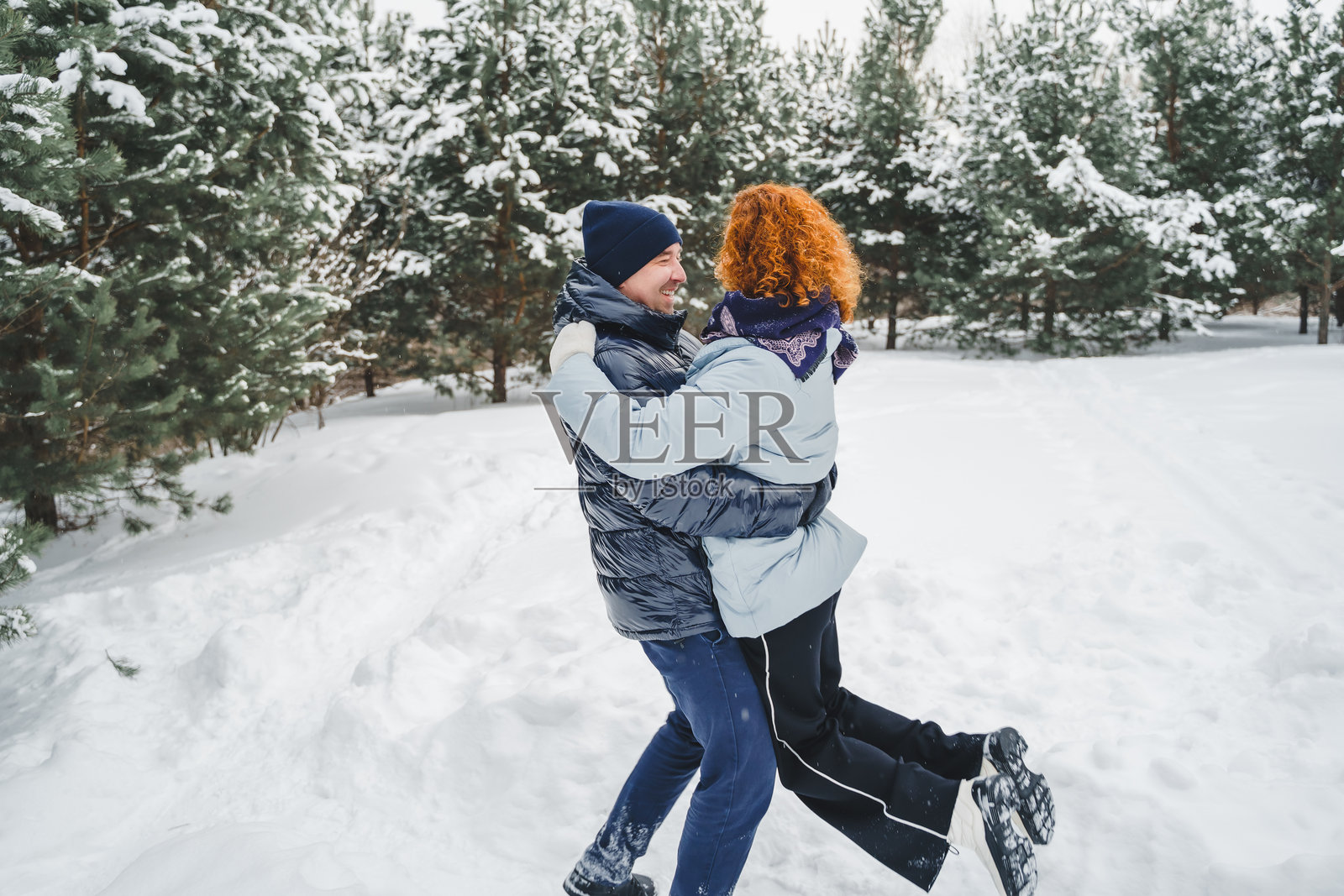 浪漫爱情故事。快乐的中年千禧夫妇，冬季雪地公园里一位红发女子。浪漫约会，亲吻，拥抱。散步，享受乐趣。时尚的蓝色衣服，夹克，帽子。照片摄影图片